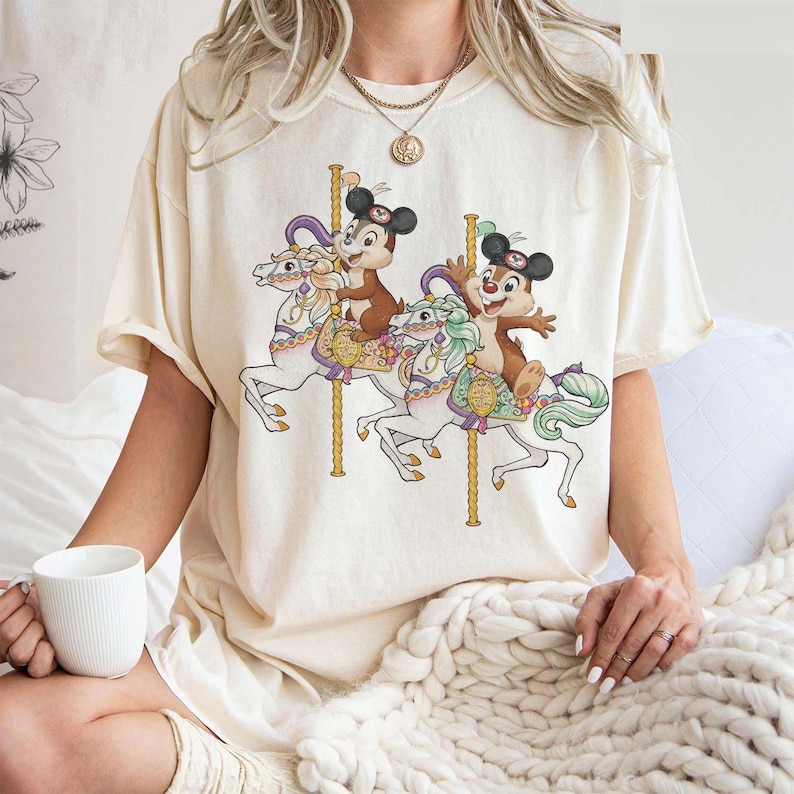May include: Cream-colored t-shirt featuring a carousel scene with cartoon characters. The design shows two chipmunk-like characters riding white carousel horses with gold poles. The horses have colorful saddles and manes. The shirt has short sleeves.