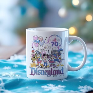 Puede incluir: Taza de cer&aacute;mica blanca con una colorida ilustraci&oacute;n de personajes de Disney, globos y fuegos artificiales. La taza conmemora el 70 aniversario de Disneyland, con el texto "Magical Year Disneyland 1955-2025". La taza tiene un asa curva.