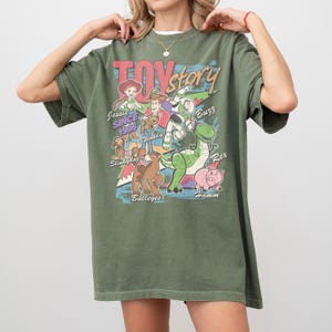 Puede incluir: Camiseta verde oliva con la frase "Toy Story" en una fuente retro, con ilustraciones de personajes como Woody, Buzz Lightyear y Rex. La camiseta tambi&eacute;n incluye el texto "Jessie Since 1995".