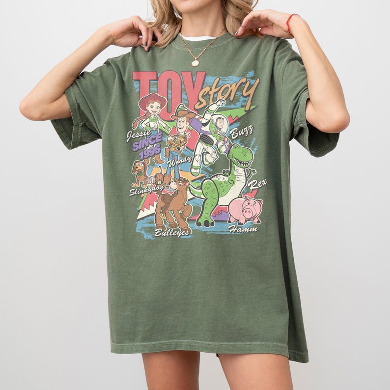 Puede incluir: Camiseta verde oliva con la frase "Toy Story" en una fuente retro, con ilustraciones de personajes como Woody, Buzz Lightyear y Rex. La camiseta tambi&eacute;n incluye el texto "Jessie Since 1995".