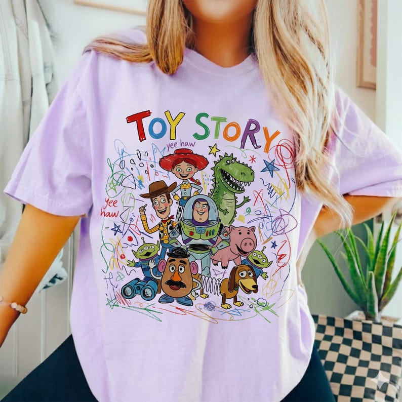 May include: A light purple t-shirt featuring a colorful graphic of Toy Story characters. The words "Toy Story" are written in a playful font above the characters. The design includes Woody, Buzz Lightyear, and other characters from the movie.