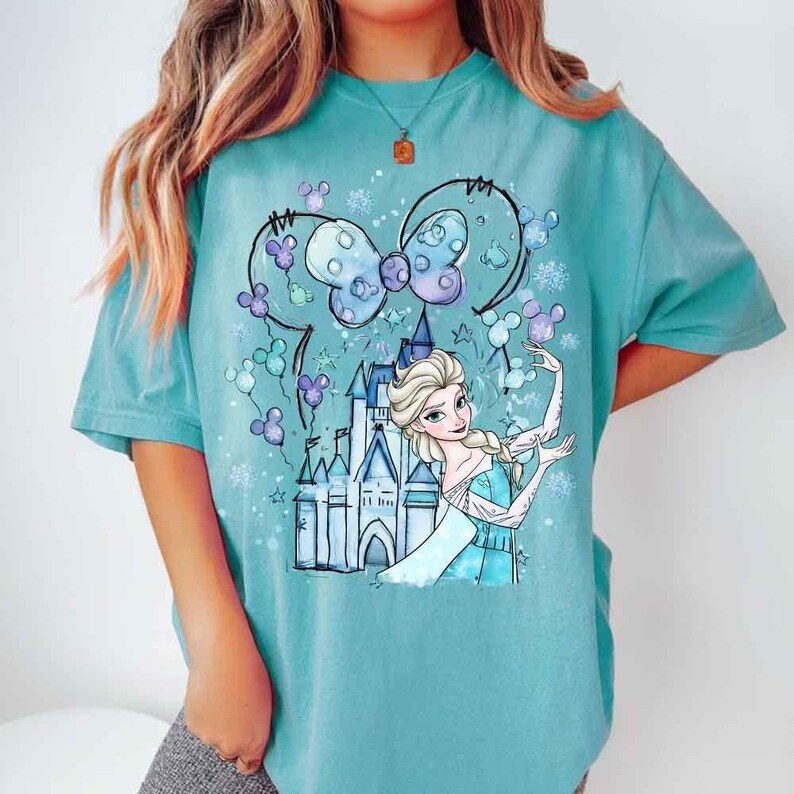 Pode incluir: Camiseta turquesa com um desenho de Elsa de Frozen, um castelo e um la&ccedil;o da Minnie. O design inclui flocos de neve e bal&otilde;es em forma de Mickey Mouse. A camisa tem mangas curtas e gola redonda.