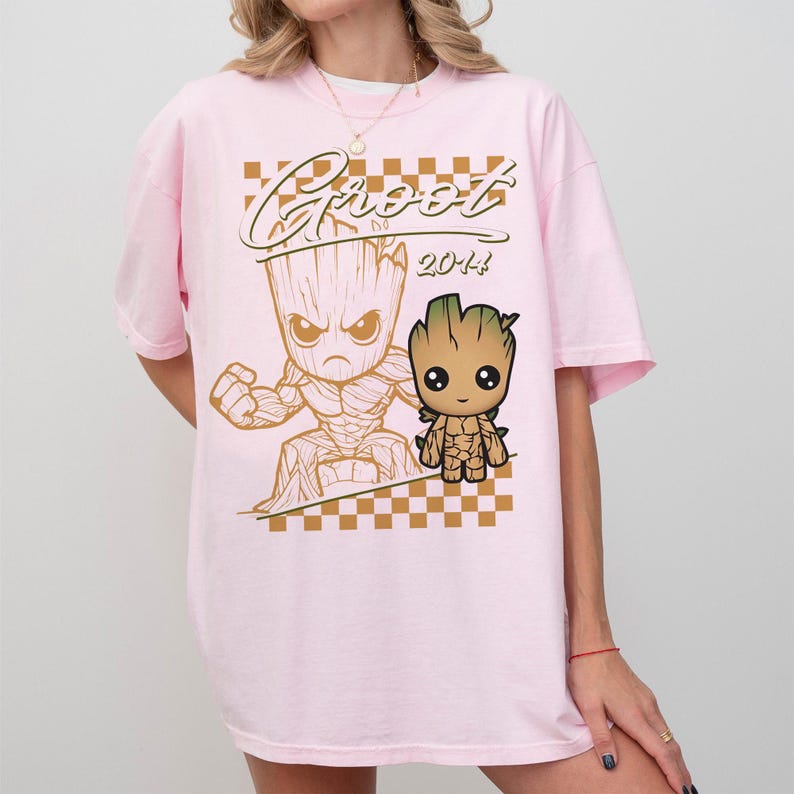 May include: A light pink t-shirt featuring a Groot graphic. The design includes the word "Groot" in a stylized font, along with two Groot illustrations and the year "2014". The shirt has short sleeves and a crew neck.