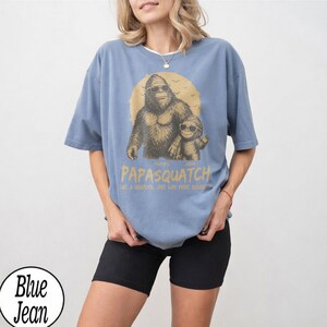 May include: A light blue t-shirt featuring a graphic of a Bigfoot with sunglasses and a smaller Bigfoot. The text on the shirt reads "Papasquatch" and "Like a Grandpa, Just Way More Squatchy."
