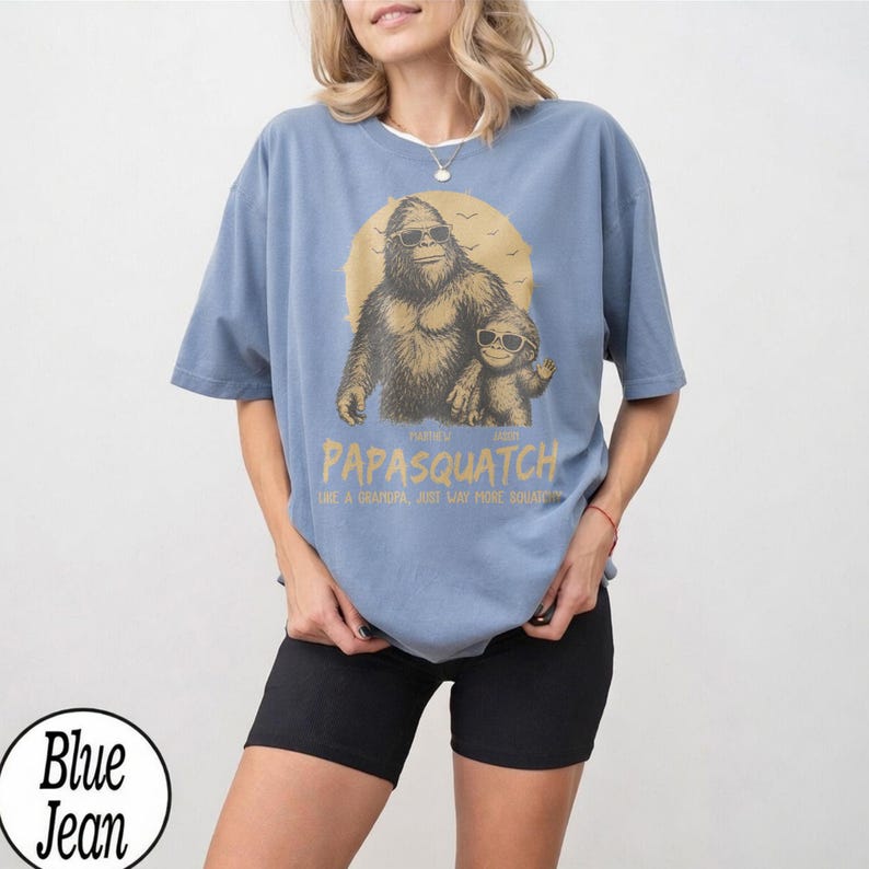 May include: A light blue t-shirt featuring a graphic of a Bigfoot with sunglasses and a smaller Bigfoot. The text on the shirt reads "Papasquatch" and "Like a Grandpa, Just Way More Squatchy."