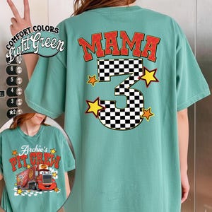 May include: Light green t-shirt with the word "MAMA" in red block letters above a large number "3" with a checkered pattern. The number is flanked by yellow stars. The shirt also features the text "Archie's Pit Crew" with a graphic of a red truck.