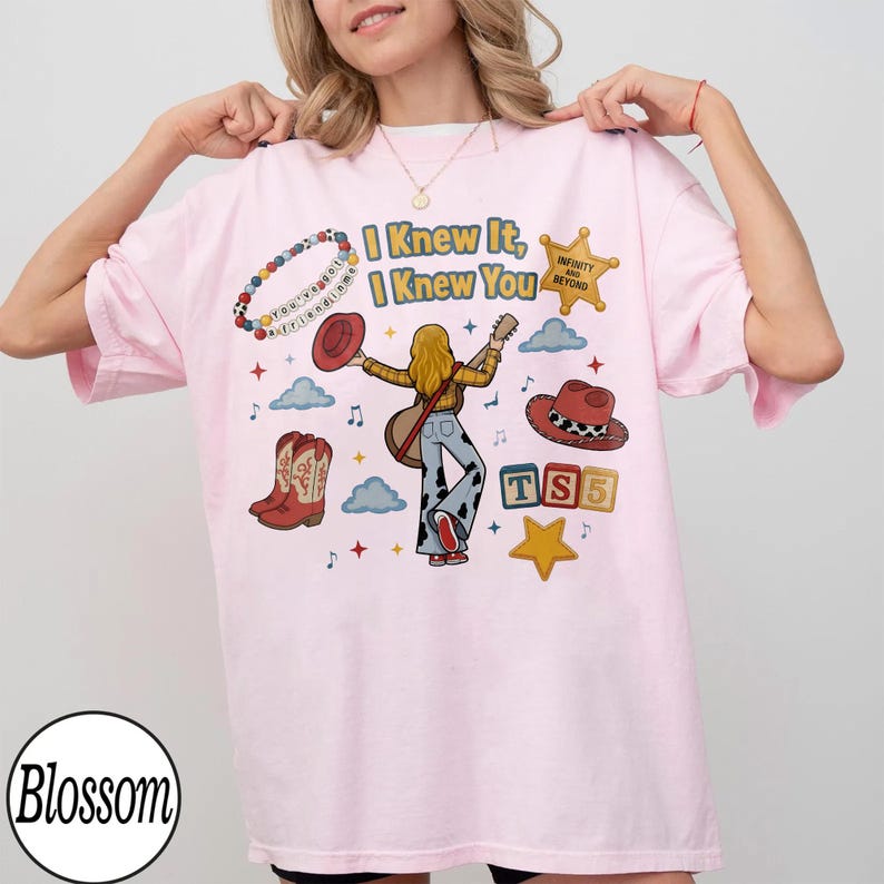 May include: A light pink t-shirt featuring a graphic print with the text I Knew It, I Knew You. The design includes a person with a guitar, a beaded bracelet, a sheriff star, cowboy boots, a hat, and blocks with the letters TS5.