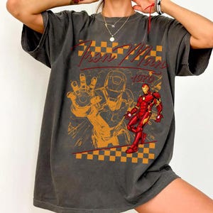 May include: Dark gray t-shirt featuring a graphic of Iron Man in red and gold, with a stylized orange illustration of Iron Man's hand and helmet. The text "Iron Man 1970" is in red script.
