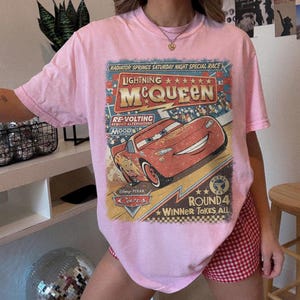 May include: A light pink t-shirt featuring a graphic print of the character Lightning McQueen from the Disney Pixar movie Cars. The design includes the text Radiator Springs Saturday Night Special Race, Re-Volting, and Round 4 Winner Takes All.