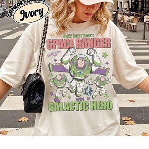 May include: Ivory-colored t-shirt featuring Buzz Lightyear from Toy Story. The design includes the text "BUZZ LIGHTYEAR'S SPACE RANGER" and "GALACTIC HERO" with images of the character. The shirt also has the words "COMFORT COLORS" and "Ivory" printed on it.