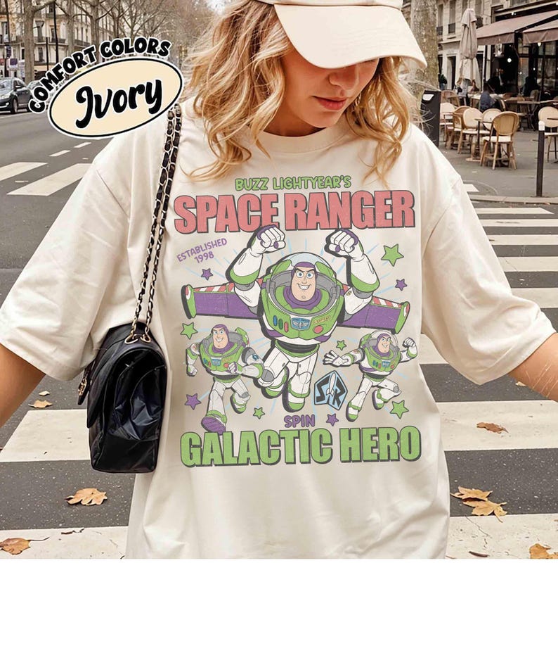 May include: Ivory-colored t-shirt featuring Buzz Lightyear from Toy Story. The design includes the text "BUZZ LIGHTYEAR'S SPACE RANGER" and "GALACTIC HERO" with images of the character. The shirt also has the words "COMFORT COLORS" and "Ivory" printed on it.