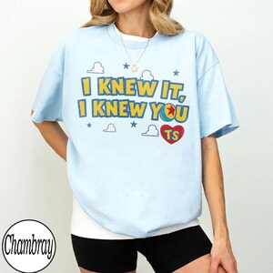 Pu&ograve; includere: Una t-shirt a maniche corte color azzurro con la scritta gialla e blu "I KNEW IT, I KNEW YOU", un cuore rosso contenente le lettere "TS" e l'icona di una palla, circondata da illustrazioni di nuvole bianche e stelle blu.