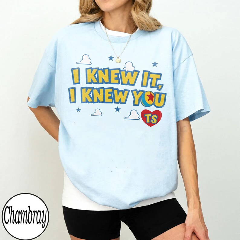 Pu&ograve; includere: Una t-shirt a maniche corte color azzurro con la scritta gialla e blu "I KNEW IT, I KNEW YOU", un cuore rosso contenente le lettere "TS" e l'icona di una palla, circondata da illustrazioni di nuvole bianche e stelle blu.