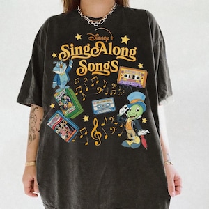 May include: Dark gray t-shirt featuring the text "Disney+ Sing Along Songs" with cartoon characters and musical notes. The design includes images of a cassette tape, Jiminy Cricket, and other Disney characters.