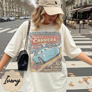 May include: An ivory short-sleeve t-shirt featuring a graphic print of the character Sally Carrera from the movie Cars. The design includes the text Sally Carrera, Radiator Springs Saturday Night Special Race, and Round 4 Winner Takes All.