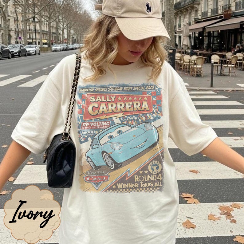 May include: An ivory short-sleeve t-shirt featuring a graphic print of the character Sally Carrera from the movie Cars. The design includes the text Sally Carrera, Radiator Springs Saturday Night Special Race, and Round 4 Winner Takes All.