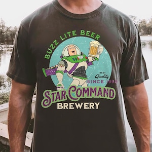 May include: Dark gray t-shirt featuring a cartoon of Buzz Lightyear holding a beer mug. The design includes the text "BUZZ LITE BEER" and "STAR COMMAND BREWERY" in green and white. The shirt has a vintage look.