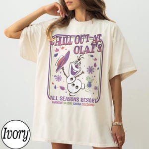 May include: Ivory-colored t-shirt featuring a graphic of Olaf from Frozen, with the text "Chill Out at Olaf's All Seasons Resort." The design includes a hat, snowflakes, and a cocktail. The shirt is a relaxed fit.
