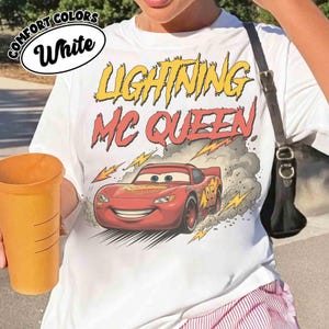 May include: White t-shirt featuring a graphic of Lightning McQueen from the Cars movie. The design includes the text "LIGHTNING MC QUEEN" in yellow and red, with a red race car and lightning bolts.