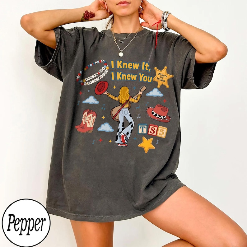 May include: A charcoal gray short-sleeve t-shirt featuring a graphic print with the text "I Knew It, I Knew You". The design includes a central figure, a cowboy hat, a pair of western boots, a star, clouds, and the text "TS5" in colorful blocks.