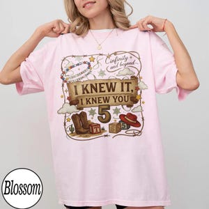 May include: A light pink t-shirt featuring a graphic print with a lasso border, a beaded necklace, and a scroll banner with the text "I KNEW IT, I KNEW YOU" above the number "5". The design includes cowboy boots, a red hat, and alphabet blocks.