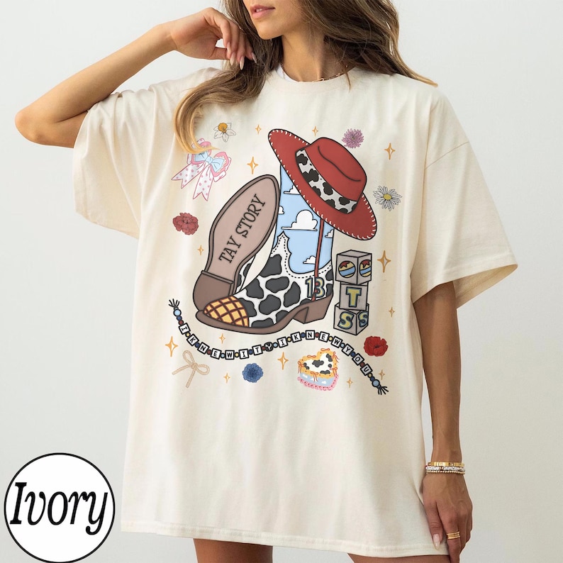 May include: An ivory t-shirt featuring a graphic print of a cowboy boot with a cow print pattern, a red hat, and the text TAY STORY. The design includes small blocks with the letters T, S, and a heart, along with floral accents and a beaded string.