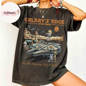 Può includere: T-shirt grigio scuro con un design di Star Wars. La grafica include il testo "Galary's Edge Star Wars" sopra un'illustrazione del Millennium Falcon, personaggi e un paesaggio. Il testo "The Most Legendary Place In The Galaxy" è sotto.