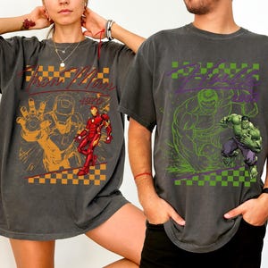 May include: Two charcoal gray t-shirts featuring comic book characters. One shirt displays "Iron Man 1970" with an orange and red graphic. The other shirt features "Hulk" with a green and purple graphic.