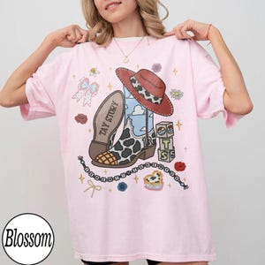 May include: A light pink t-shirt featuring a graphic print of a cowboy boot with a cow print pattern, a red hat, a blue sky background, alphabet blocks spelling TS, a bow, and text reading TAY STORY and I KNEW IT I KNEW YOU.