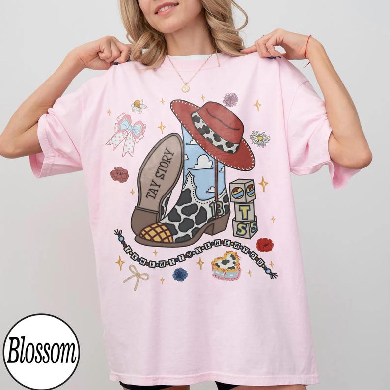 May include: A light pink t-shirt featuring a graphic print of a cowboy boot with a cow print pattern, a red hat, a blue sky background, alphabet blocks spelling TS, a bow, and text reading TAY STORY and I KNEW IT I KNEW YOU.