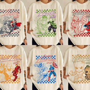 May include: Cream-colored t-shirts featuring various Marvel comic book characters. Designs include Thor, Hulk, Spider-Man, Iron Man, Captain America, and Groot, each with their name and year. The graphics are in a comic book style with a checkered pattern.