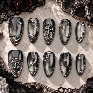 May include: A set of ten almond-shaped press-on nails in a dark charcoal gray with a metallic cat-eye finish. The nails feature raised silver-toned 3D embellishments, including ornate cross motifs, swirling liquid-like patterns, and small clear rhinestones.