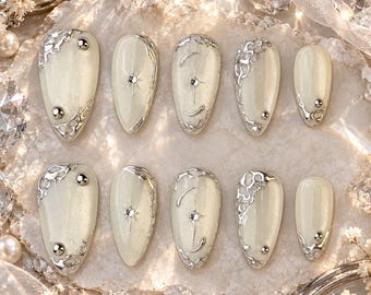 May include: A set of ten almond-shaped press-on nails in a pearlescent off-white finish. The nails feature raised silver metallic liquid-style accents, small silver studs, and delicate star-shaped engravings with tiny embedded crystals.