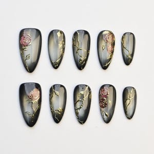 Puede incluir: Juego de diez uñas postizas de forma ovalada. Cada uña presenta una base degradada gris y negra con detalles dorados en forma de vid. Algunas uñas tienen un diseño de rosa rosa. Las uñas están dispuestas sobre una superficie blanca.