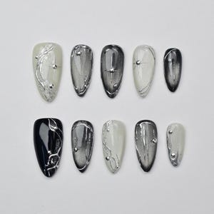 May include: A collection of ten almond-shaped false nails. The nails display diverse designs, including solid black, gradient grey, and off-white. Each nail is decorated with silver embellishments, such as lines and small studs.