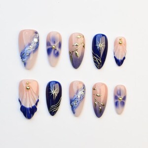 Può includere: Un set di dieci unghie finte con vari design. Le unghie presentano una miscela di colori, tra cui blu navy, rosa chiaro e oro. Alcune unghie hanno motivi floreali o astratti, altre dettagli in foglia d'oro.