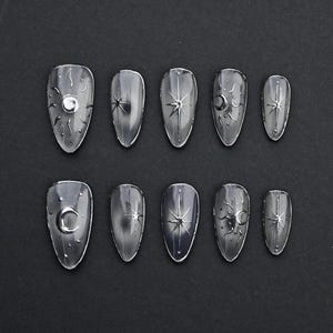May include: A set of silver-toned, press-on nails. Each nail features celestial designs, including suns, stars, and crescent moons. The nails are arranged on a black surface, showcasing their metallic sheen and intricate detailing.