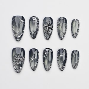 Pode incluir: Um conjunto de dez unhas postiças em vários tamanhos. Cada unha é de cor cinza escuro com enfeites prateados. Algumas unhas apresentam um desenho de cruz, enquanto outras têm padrões abstratos. As unhas têm um acabamento brilhante.