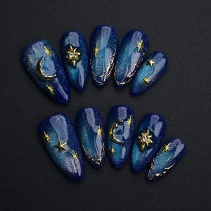 May include: A set of ten almond-shaped artificial nails. The nails are painted a deep blue with a glittery finish. Each nail features celestial-themed embellishments, including gold stars, moons, and decorative accents.