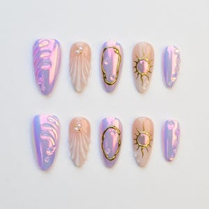 Puede incluir: Un conjunto de diez uñas postizas iridiscentes con diversos diseños. Las uñas presentan detalles rosas, morados y dorados, incluyendo espirales, soles y contornos dorados. Algunas uñas están adornadas con pequeños adornos de perlas.