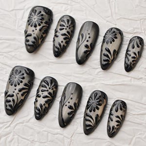 Op de afbeelding: Een set van tien donkergrijze persnagels met een glanzende afwerking. Elke nagel heeft een verhoogd zwart bloemmotief. De nagels zijn gerangschikt op een witte, getextureerde ondergrond, die de details van de nail art benadrukt.