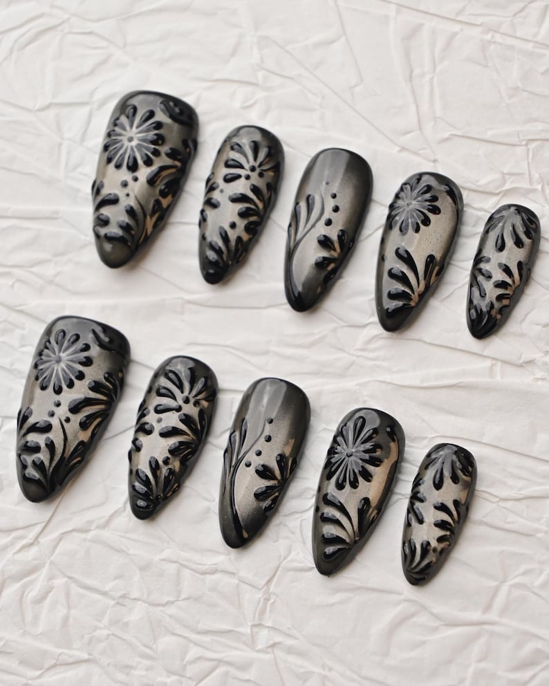 Op de afbeelding: Een set van tien donkergrijze persnagels met een glanzende afwerking. Elke nagel heeft een verhoogd zwart bloemmotief. De nagels zijn gerangschikt op een witte, getextureerde ondergrond, die de details van de nail art benadrukt.