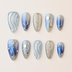 Puede incluir: Un conjunto de diez uñas artificiales con varios diseños. Las uñas presentan una paleta de colores plateado, azul y gris. Algunas uñas tienen un diseño plateado ondulado, mientras que otras tienen adornos de estrellas y gemas. Las uñas son de forma ovalada.