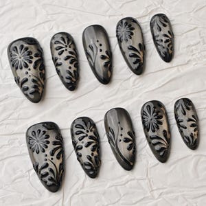 Op de afbeelding: Een set van tien grijze en zwarte nepnagels. Elke nagel heeft een glanzende afwerking en een verhoogd, bloemmotief in zwart. De nagels zijn ovaal van vorm en gerangschikt op een wit, getextureerd oppervlak.