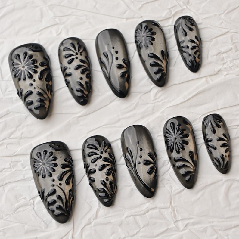 Op de afbeelding: Een set van tien grijze en zwarte nepnagels. Elke nagel heeft een glanzende afwerking en een verhoogd, bloemmotief in zwart. De nagels zijn ovaal van vorm en gerangschikt op een wit, getextureerd oppervlak.