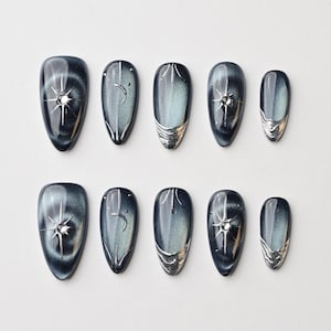 May include: A set of ten oval-shaped false nails. Each nail features a dark blue and silver design with celestial elements, including stars and crescent moons. Some nails have a silver metallic accent at the base.