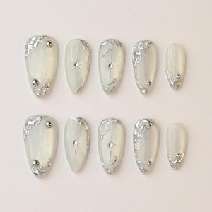 May include: A set of ten almond-shaped, press-on nails. Each nail is a pearlescent white with silver embellishments. Some nails feature silver swirls, while others have a star or crescent moon design with a central gem.