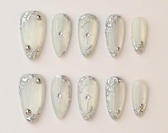 May include: A set of ten almond-shaped, press-on nails. Each nail is a pearlescent white with silver embellishments. Some nails feature silver swirls, while others have a star or crescent moon design with a central gem.