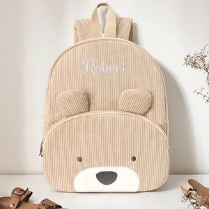 May include: A tan corduroy backpack with a bear face design. The backpack features embroidered text "Robert" in white, bear ears, a zippered front pocket, and a top handle. The bag is approximately 12 inches tall.
