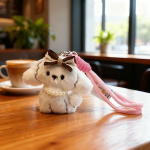 Może przedstawiać: Brelok do kluczy z białym pluszowym psem z brązową kokardą i koronką, perłowym naszyjnikiem i różową smyczą. Pies ma czarne oczy i mały czarny nos. Brelok leży na drewnianym stole.
